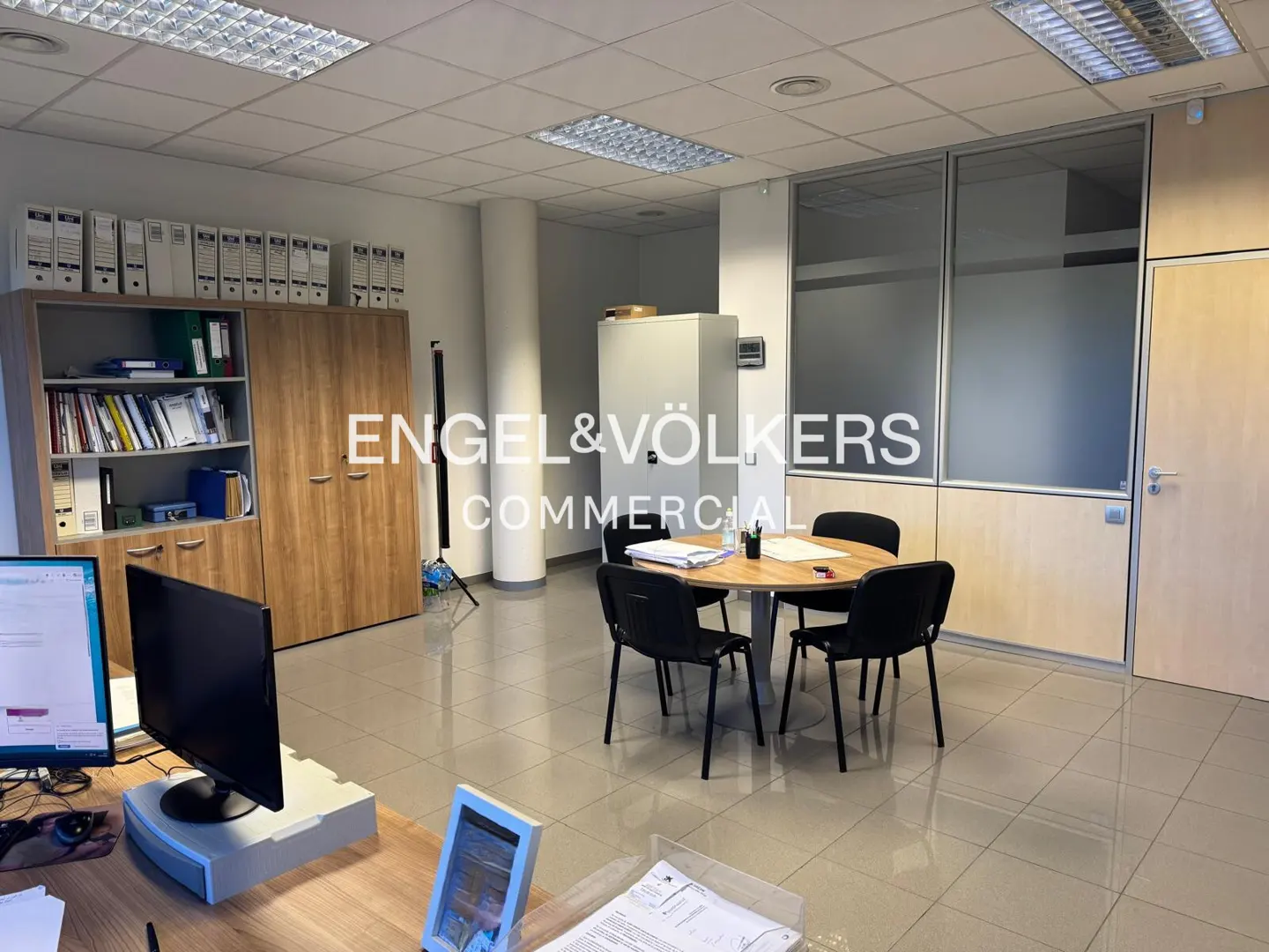 Bright office space with a round table, black chairs, filing cabinets, and Engel & Völkers Commercial logo on a glass wall.