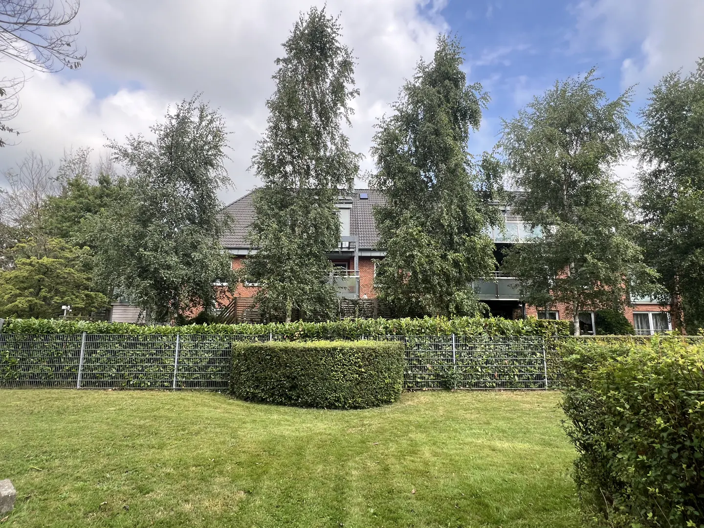 Exterior view of a brick apartment building with green lawn, trees, and a hedge fence.