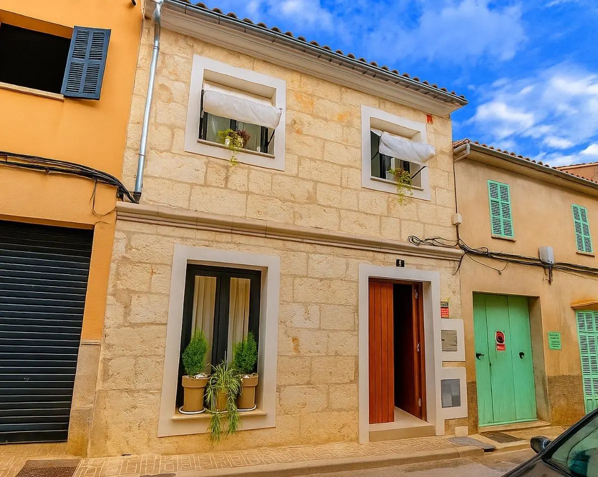 Two-story stone house with a wooden door, potted plants in windows, and adjacent yellow and tan buildings under blue sky.