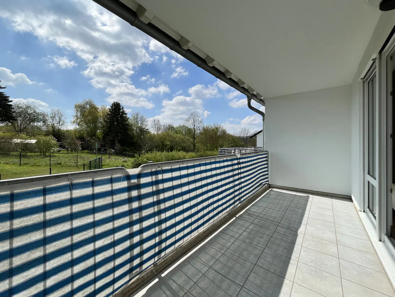 Balcony view with blue and white striped privacy screen, gray tiled floor, and a view of trees and sky.