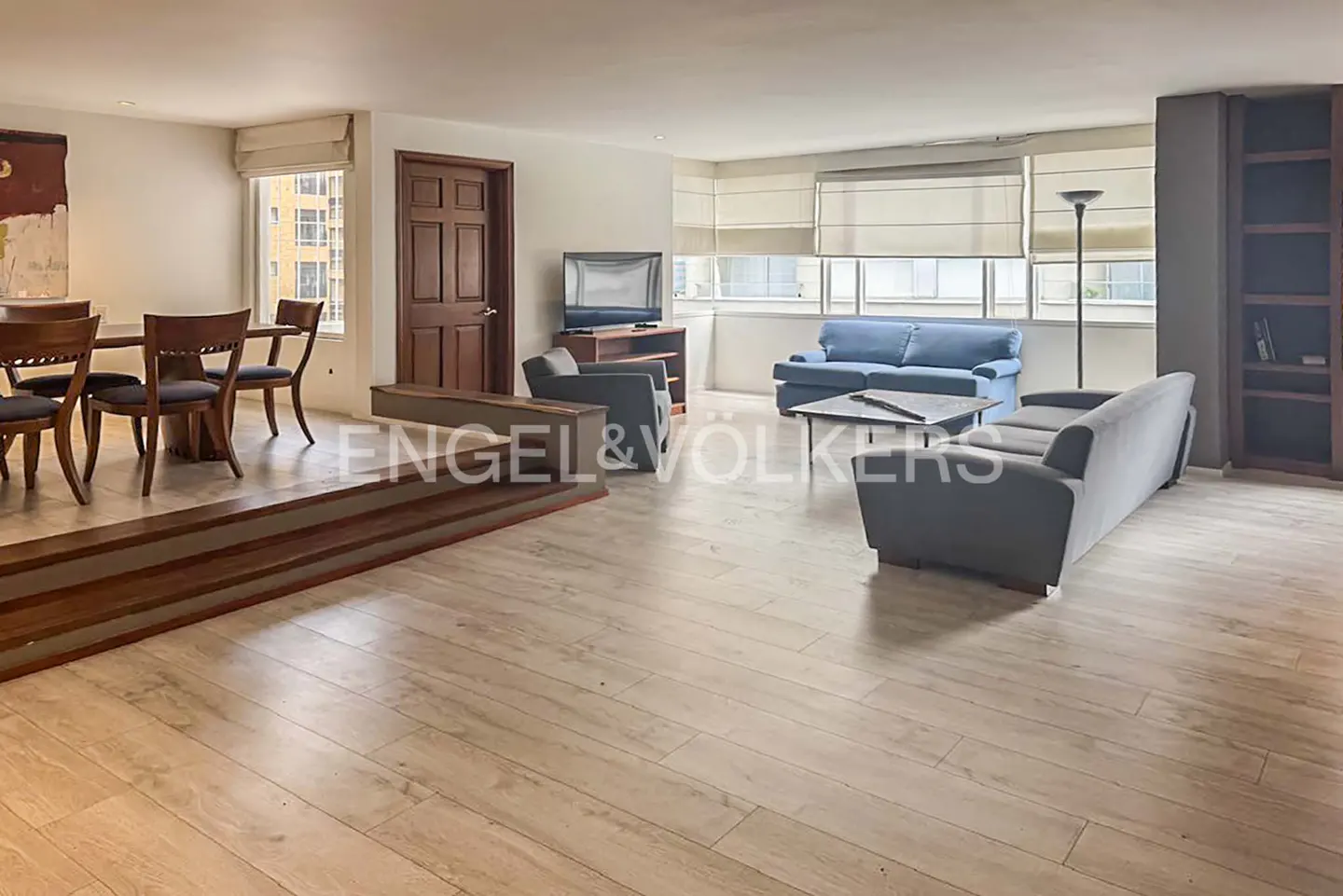 Spacious living room with light wood floors, a sunken dining area, blue sofa, grey couch, and large windows with blinds.