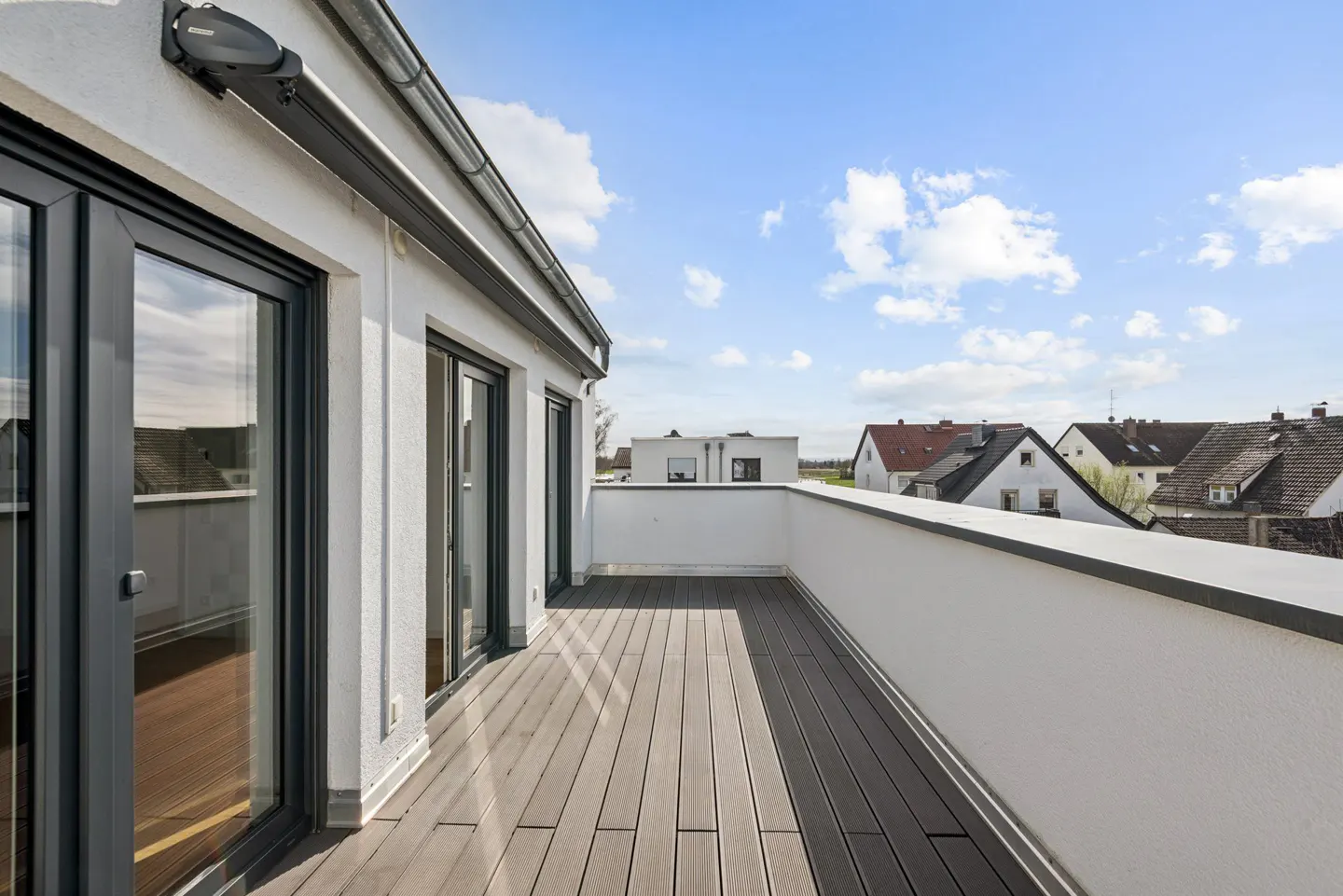 A modern balcony with gray wood flooring, white walls, and black-framed glass doors, overlooking houses under a blue sky.