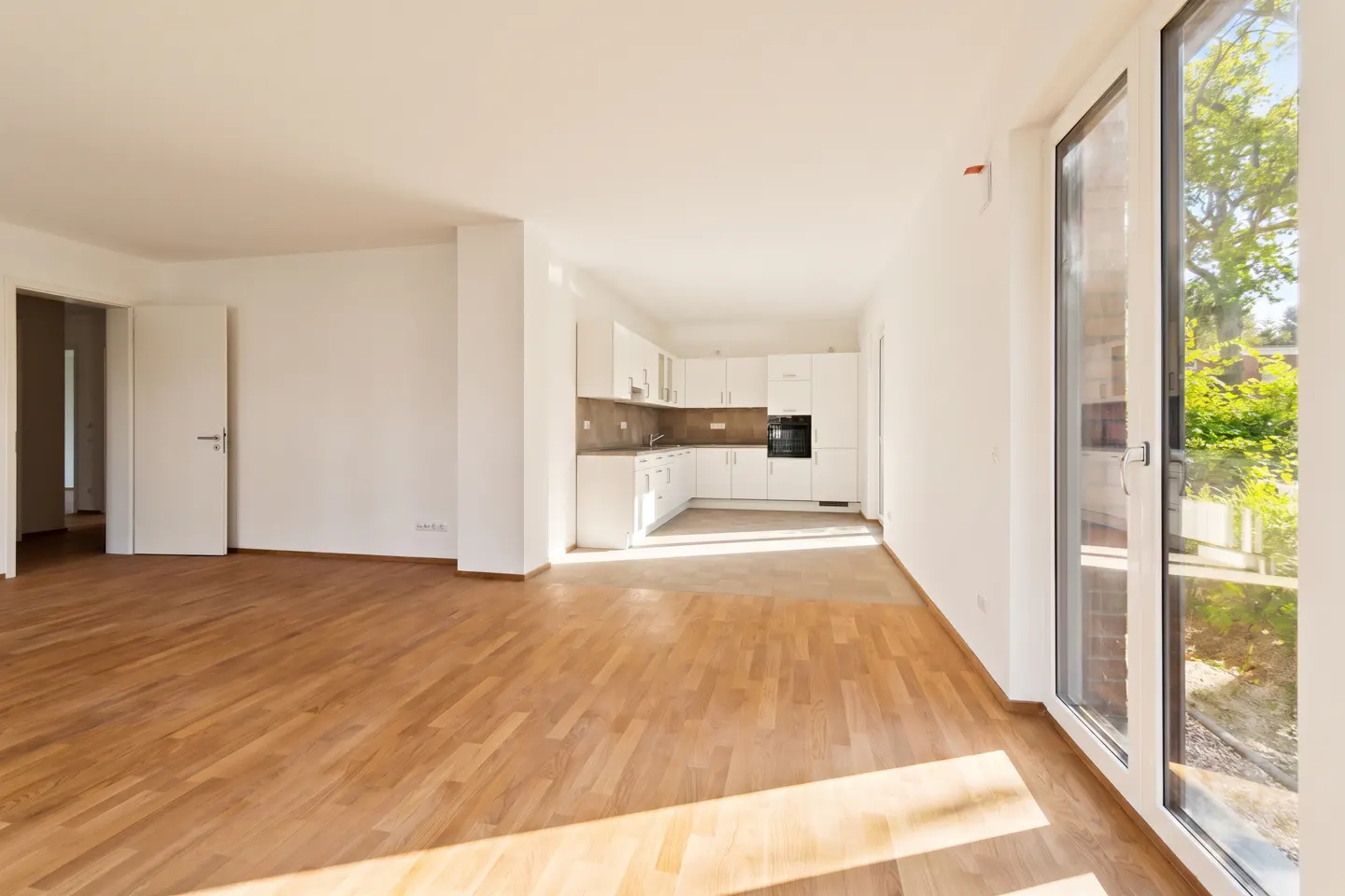 Bright, empty apartment with hardwood floors, white walls, and a white kitchen. Sunlight streams in through a glass door.