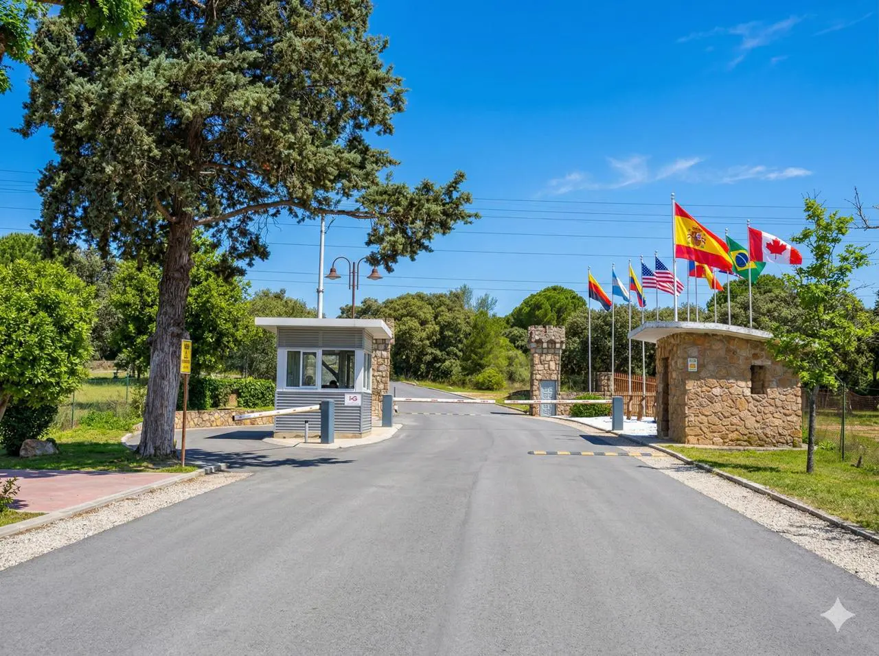 Gated entrance to a property with international flags. Guardhouses are stone and gray. The sky is blue.