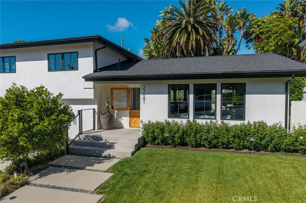 Two-story white house with black trim, dark roof, and a wooden door. Green lawn and landscaping in front. Blue sky.