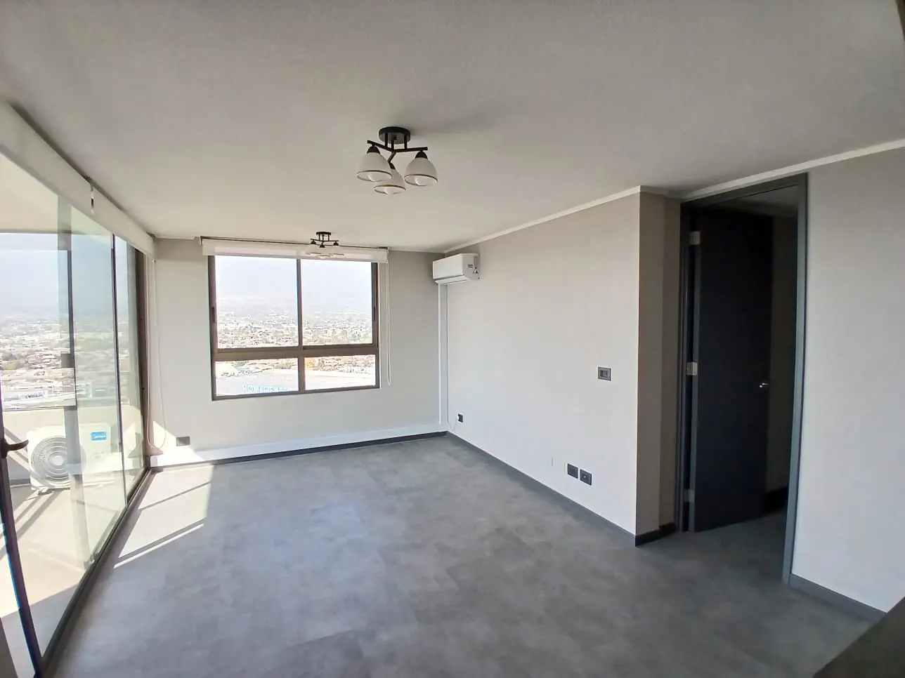 Bright, empty room with gray floors, white walls, and large windows overlooking a city and ocean view. A dark door is slightly ajar.