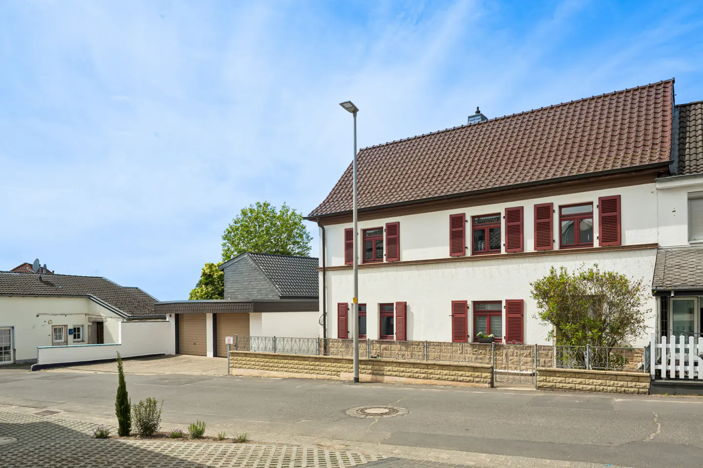 Two-story white house with brown roof and red shutters, featuring a stone base, attached garage, and a clear blue sky.
