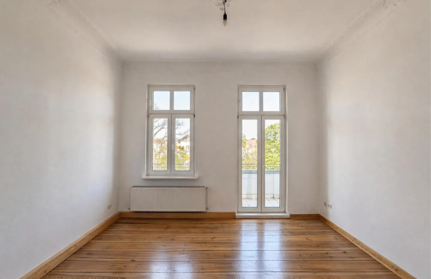 Empty room with white walls, wood floors, and two white windows. One window is a door to a balcony.
