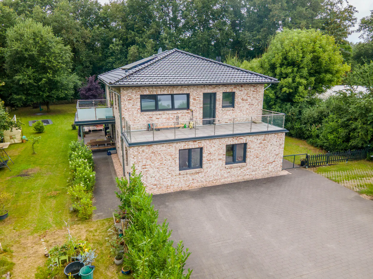 Two-story brick house with a gray roof, black framed windows, and a balcony. A paved driveway leads to the house, surrounded by green trees.