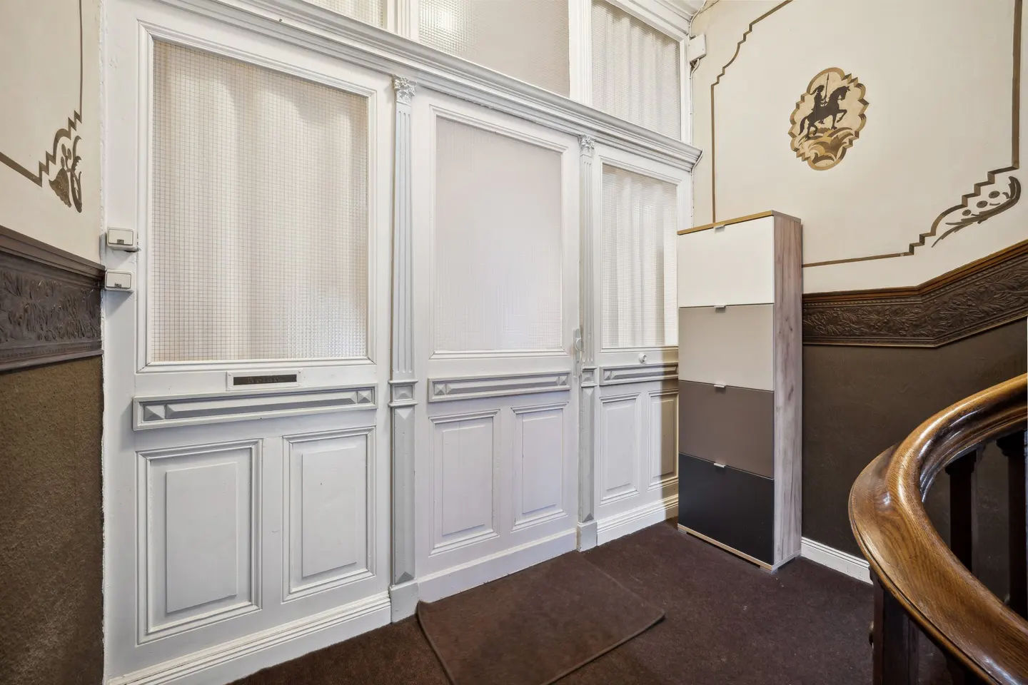 Hallway with white paneled walls, a shoe cabinet, and a brown carpet. A horse and rider design is on the wall.