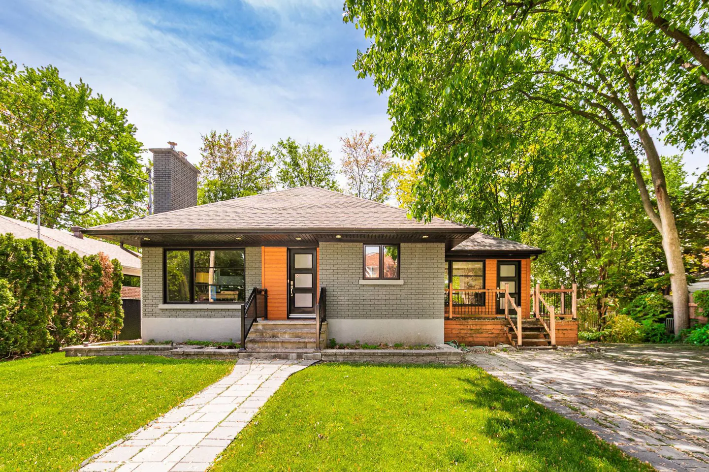 Single-story grey brick house with orange accents, a stone walkway, and a lush green lawn under a bright blue sky.