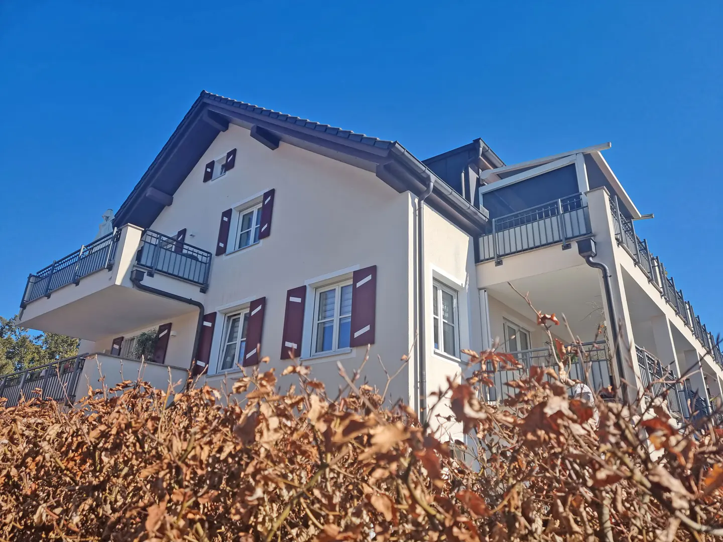 A two-story house with balconies, cream walls, and maroon shutters under a clear blue sky. A brown hedge is in the foreground.