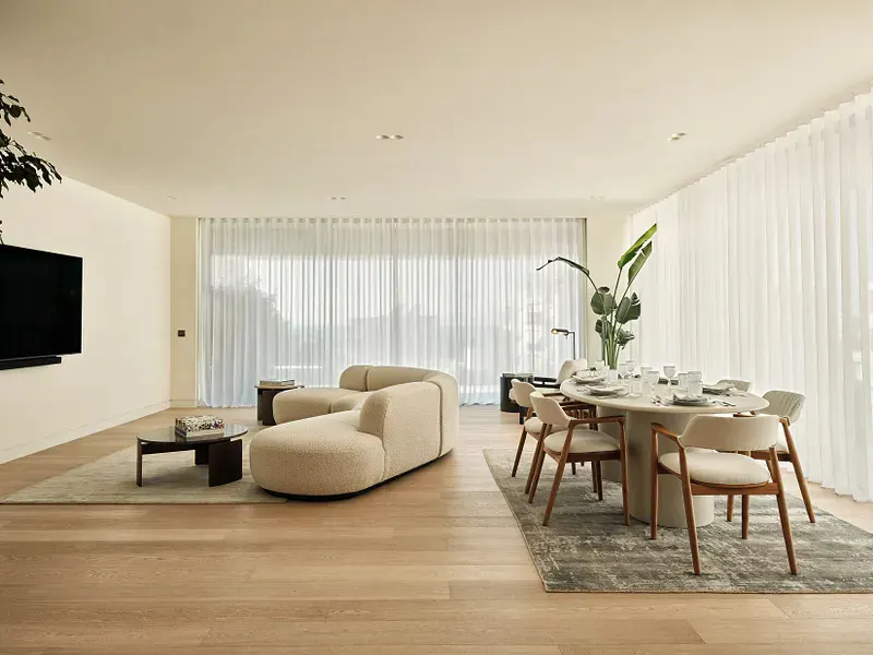 Bright, modern living and dining area with a cream sofa, oval table set for dining, and light wood floors. White curtains filter sunlight.