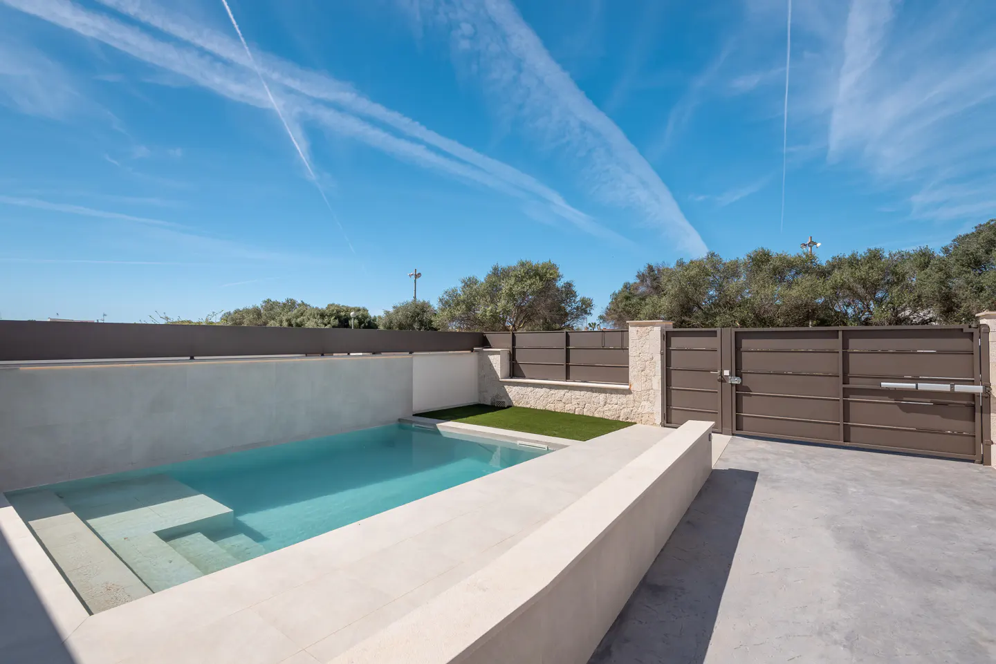 A modern, rectangular pool with steps, surrounded by a stone patio, a small patch of grass, and a brown metal gate under a blue sky.