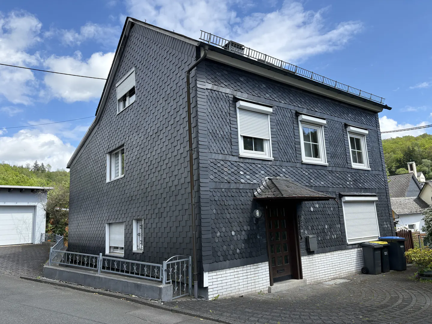Two-story house with dark gray shingle siding, white trim, and roller shutters on some windows. A small porch covers the front door.