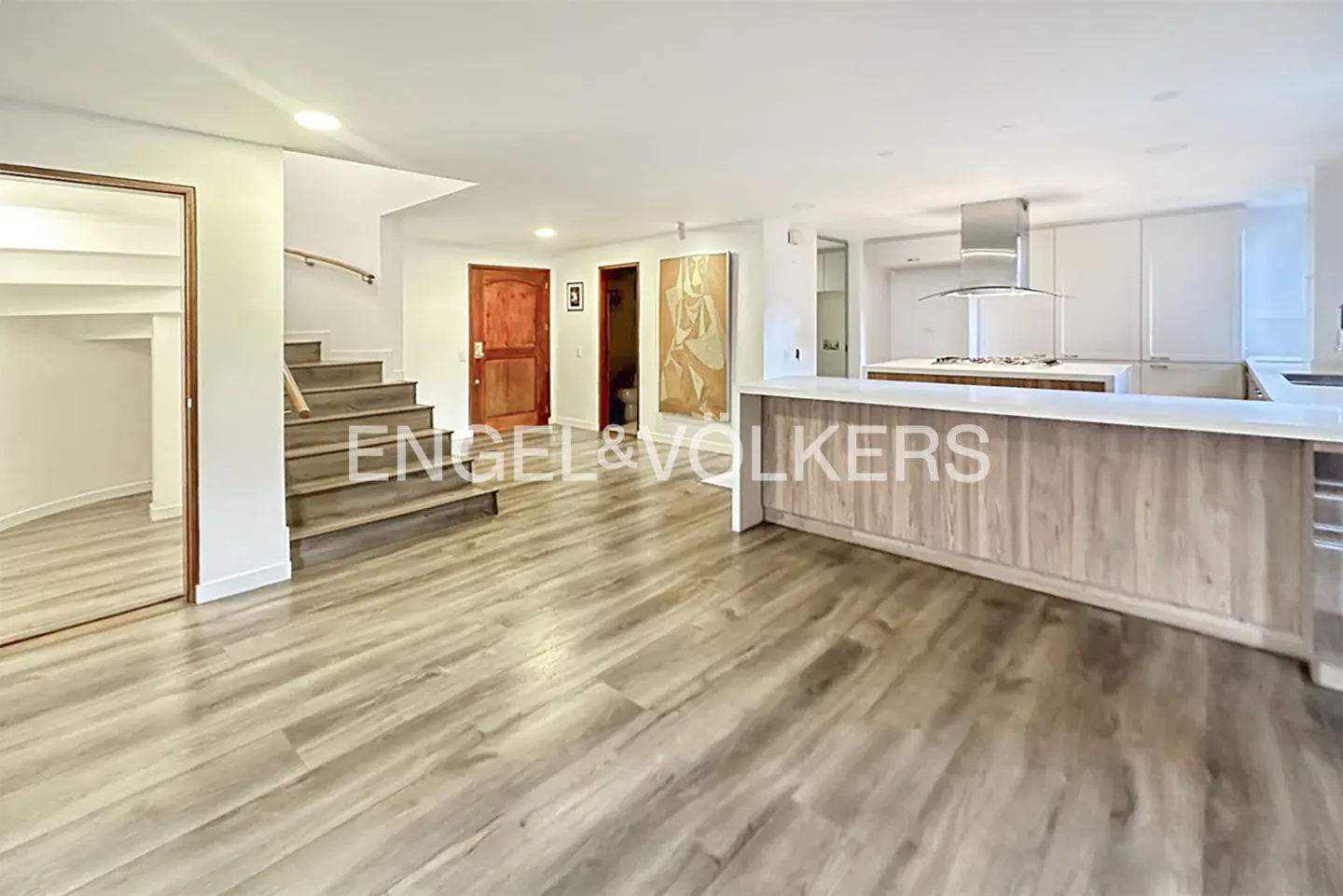 Open-concept living space with wood floors, stairs, and a kitchen island with white countertops. A wooden door and artwork are visible.