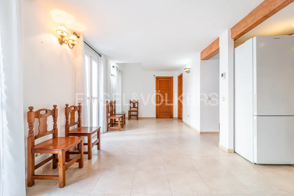 Bright, empty room with tile floor, white walls, and wooden chairs. A white refrigerator stands near a doorway. Natural light streams in.