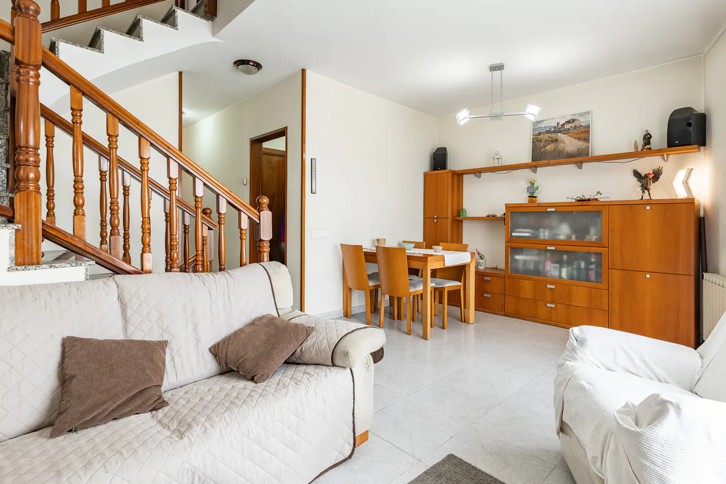 A bright living room with a white sofa, wooden staircase, dining table, and a wooden cabinet with shelves.