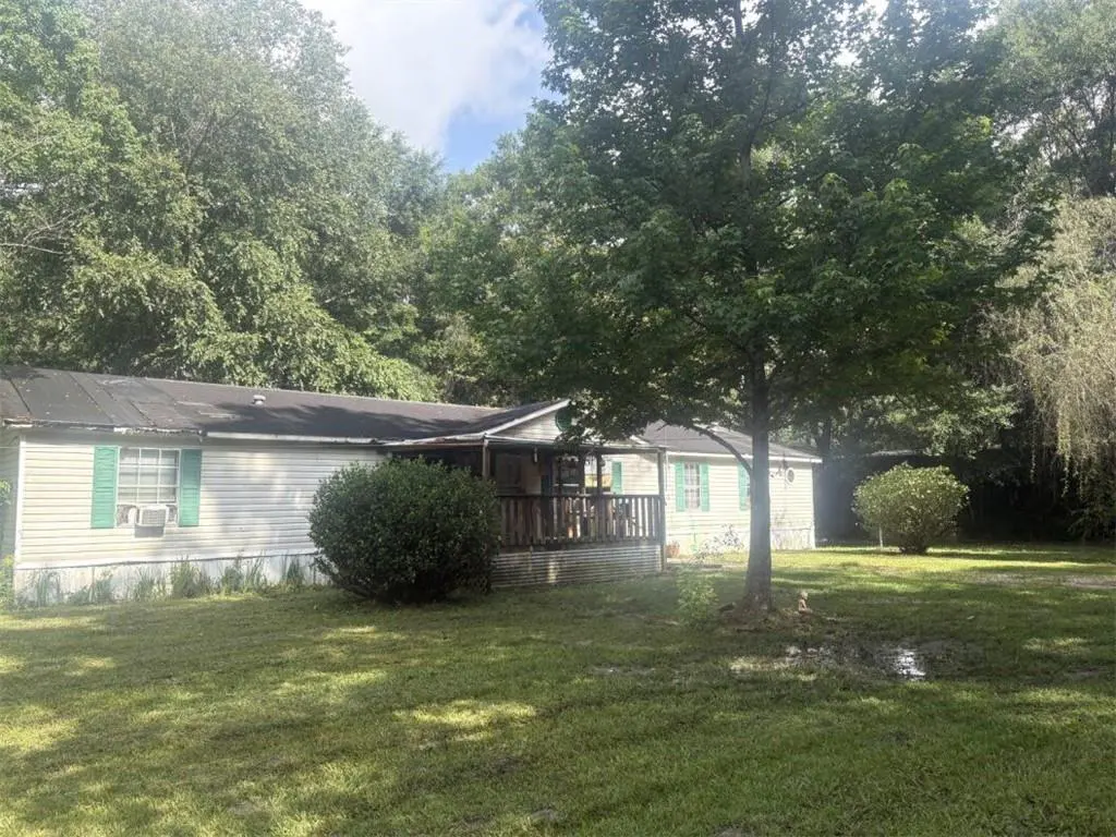 Single-story mobile home with light siding, green shutters, and a wooden front porch, set in a large, tree-filled yard.