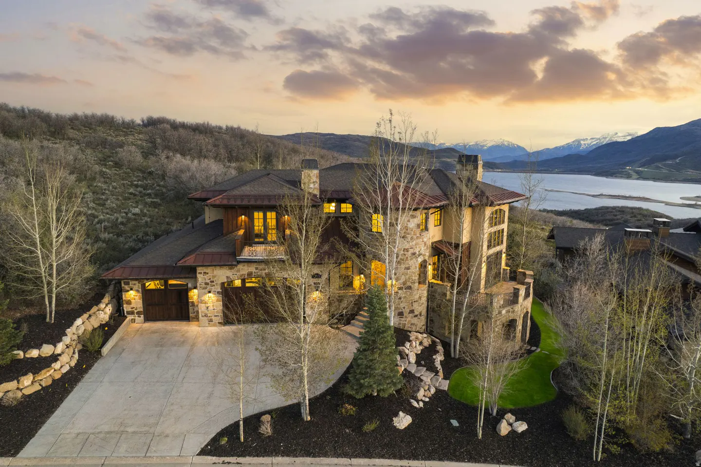 Stone house with a dark roof, a driveway, and a view of a lake and mountains at sunset.
