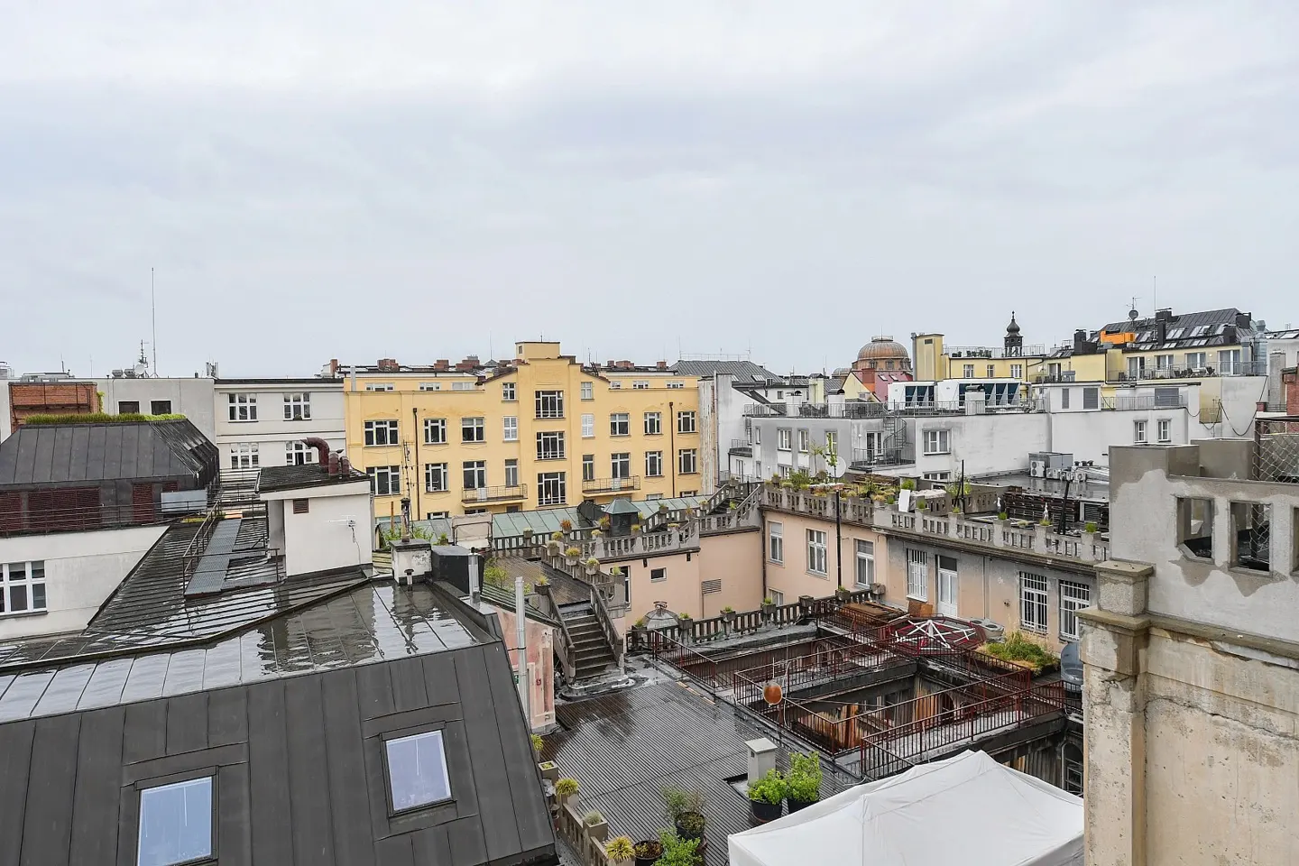 Cityscape view of rooftops with various colors and textures under a cloudy sky. Some rooftops have plants and outdoor furniture.