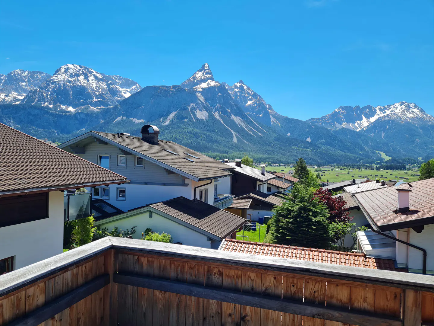 View from a wooden balcony overlooking houses and snow-capped mountains under a clear blue sky.