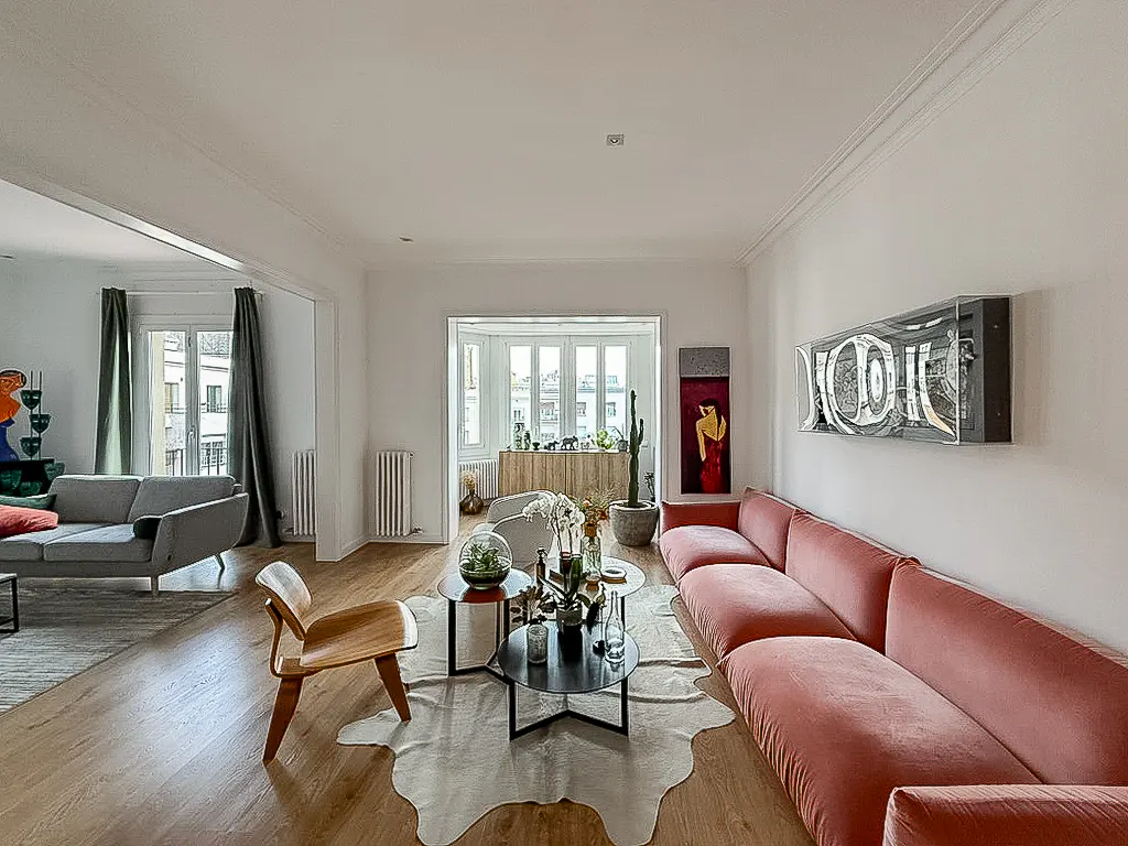Bright living room with a pink sofa, wooden floors, cowhide rug, coffee tables, and a view into an adjacent sunroom.