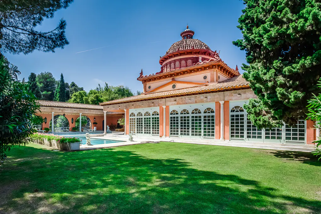 Mediterranean villa with a domed roof, large glass windows, and a swimming pool set in a lush green garden.