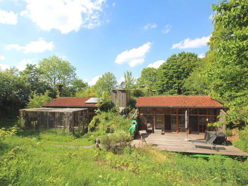 A rustic house with a red-tiled roof, wooden deck, and large outdoor animal enclosure surrounded by lush green trees.