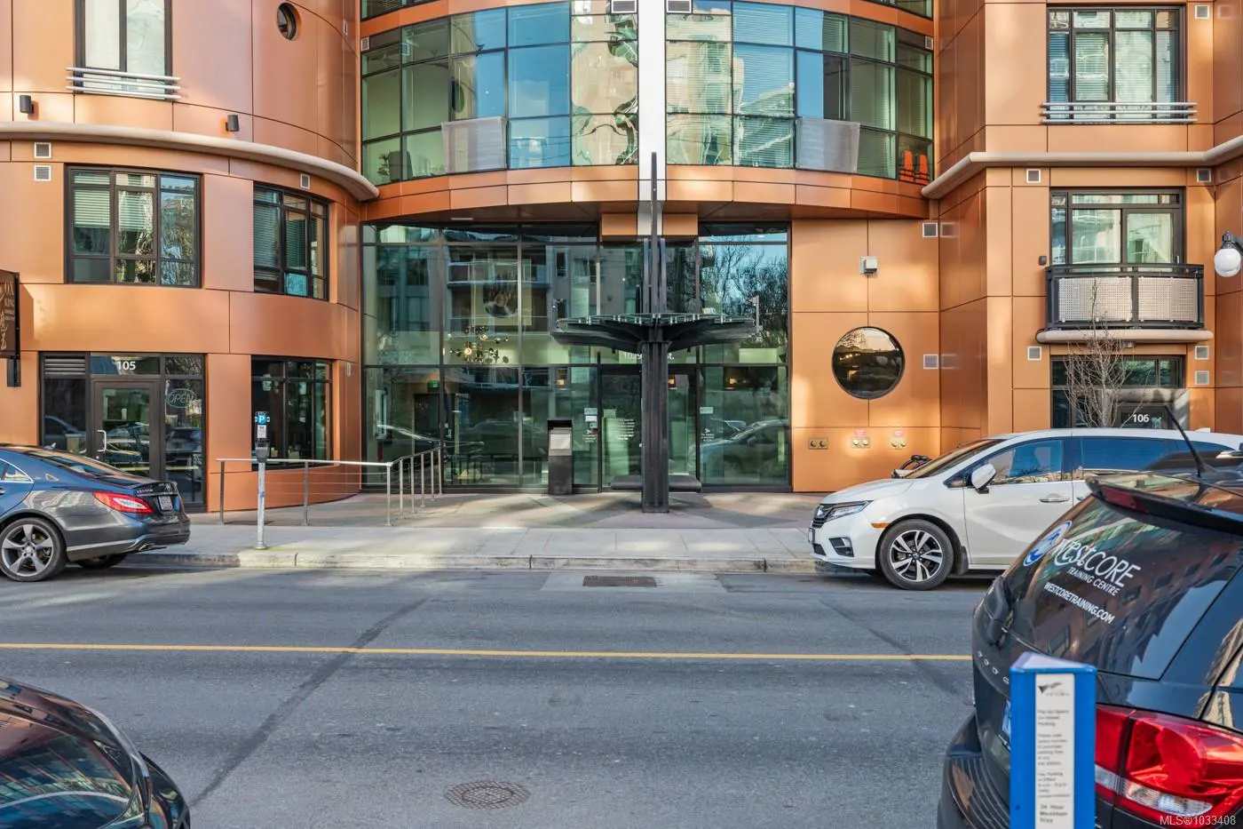 Exterior view of a modern, copper-colored building with glass windows and a unique entrance canopy. Cars are parked on the street.