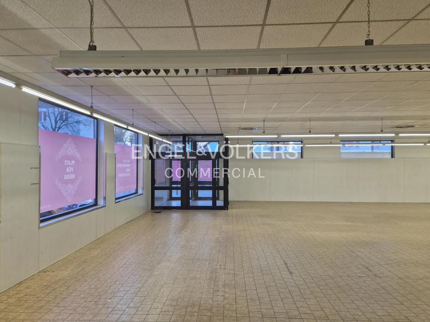 Empty commercial space with tile floors, white walls, and large windows. Engel & Volkers Commercial logo on the wall.
