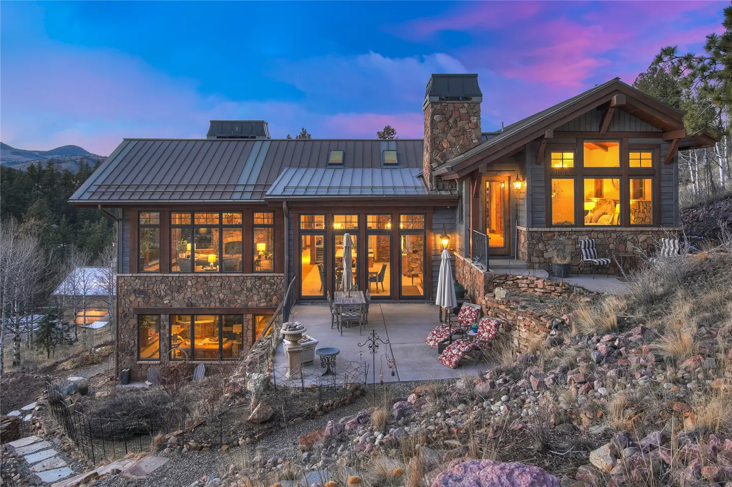 Exterior view of a stone and wood home with a metal roof, patio, and mountain backdrop at dusk.