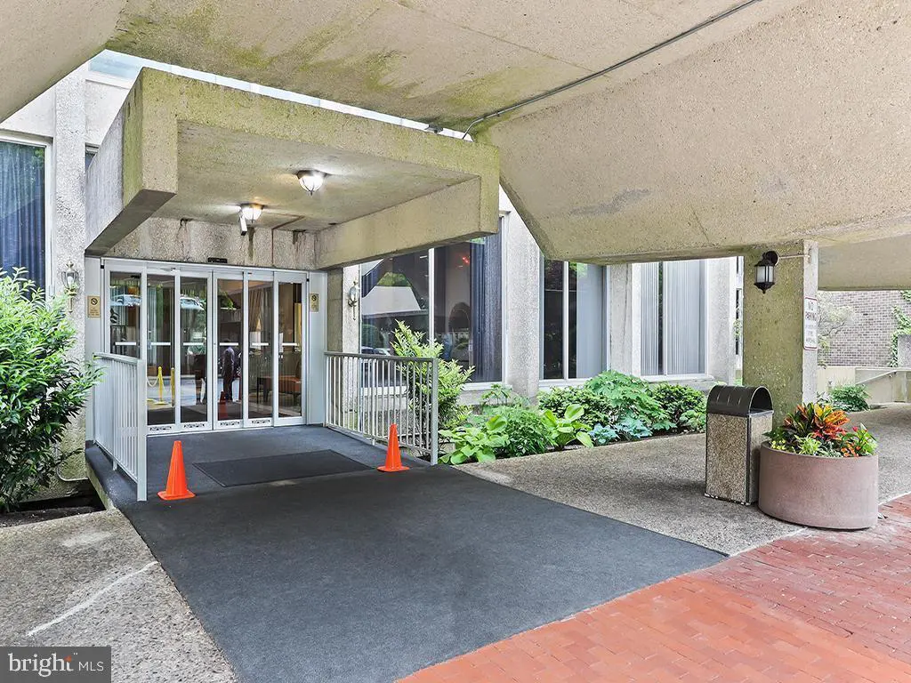 Exterior view of a building entrance with automatic doors, concrete overhang, and landscaping. Two orange cones are placed near the entrance.