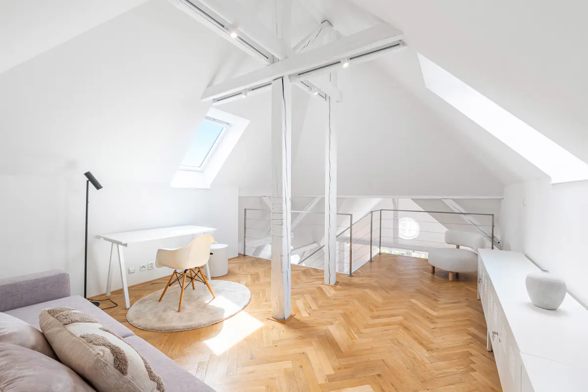 Bright attic living space with white walls, exposed beams, and a skylight. A desk, chair, sofa, and herringbone wood floors furnish the room.
