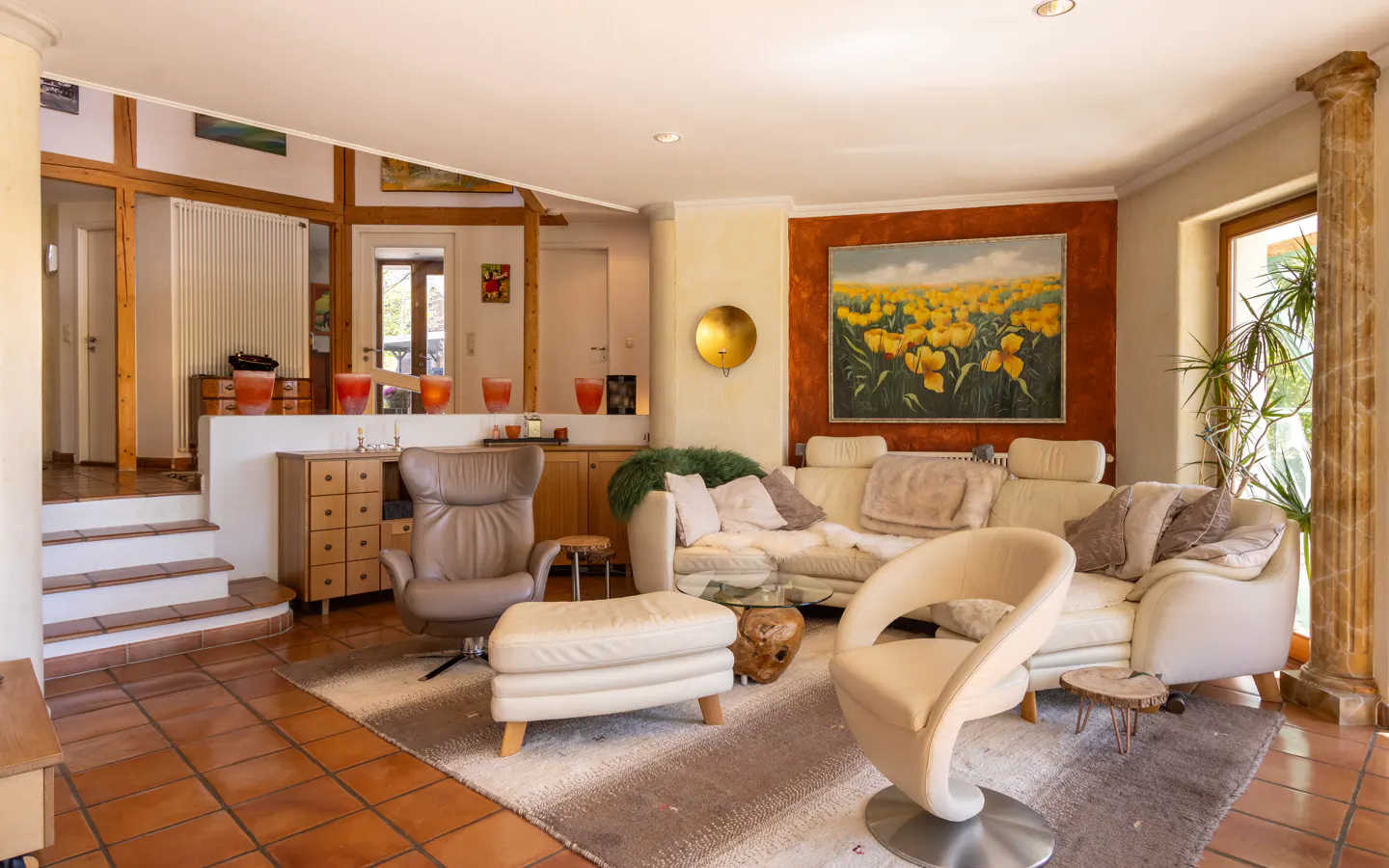 A living room with a white sofa, beige chairs, and a painting of yellow flowers. The floor is tiled in terracotta.