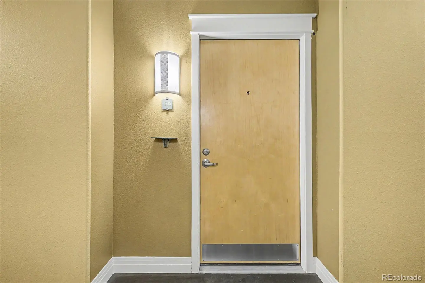 Apartment hallway with a light wood door, white trim, and a silver door handle. A sconce light is on the yellow wall.