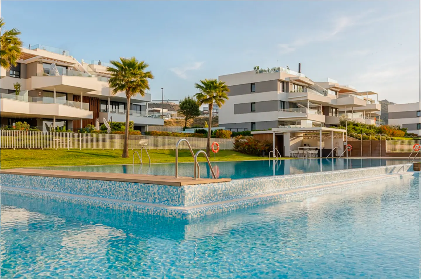 A blue tiled pool with metal ladder, surrounded by green grass and modern white buildings with palm trees under a clear sky.