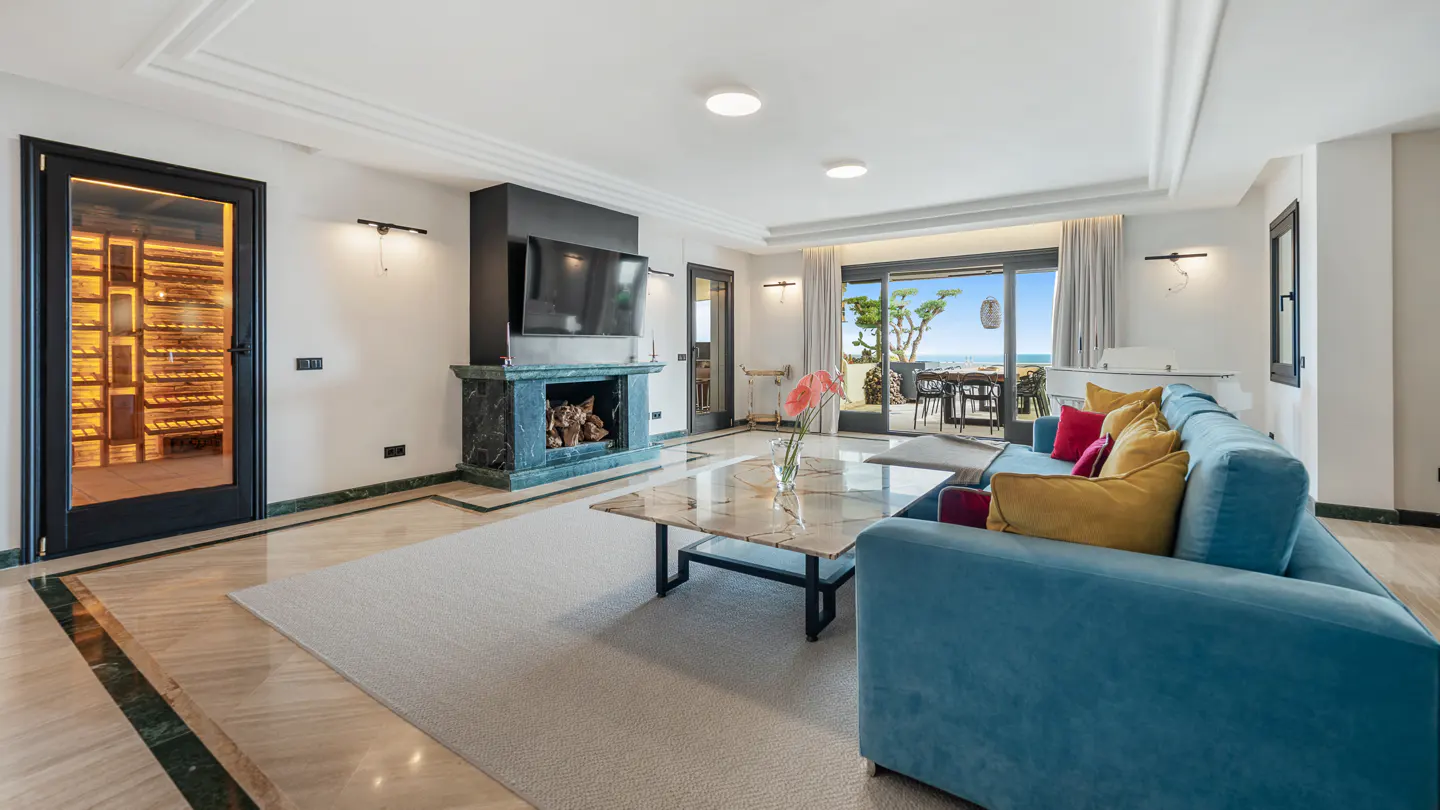Bright living room with blue sofa, marble fireplace, and wine cellar. Ocean view through sliding glass doors.