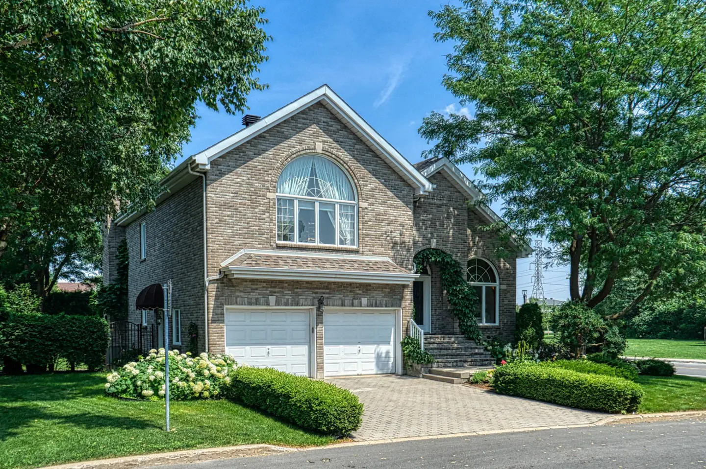 Two-story brick house with white garage doors, arched window, and manicured lawn under a blue sky.