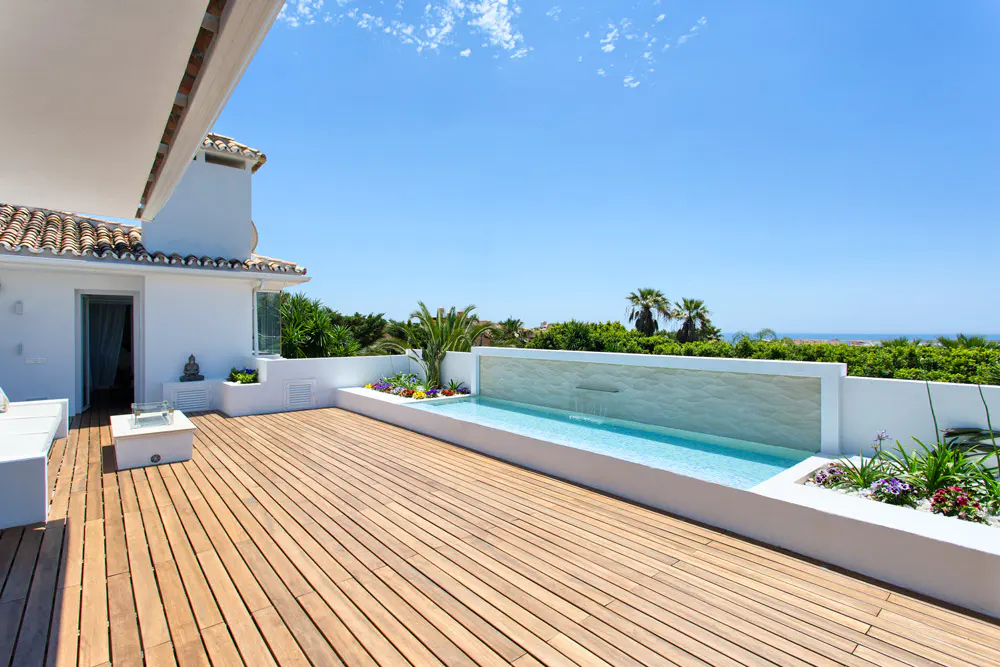 Luxury rooftop terrace with a wooden deck, a narrow pool with a waterfall feature, and white walls. Palm trees and blue sky in the background.