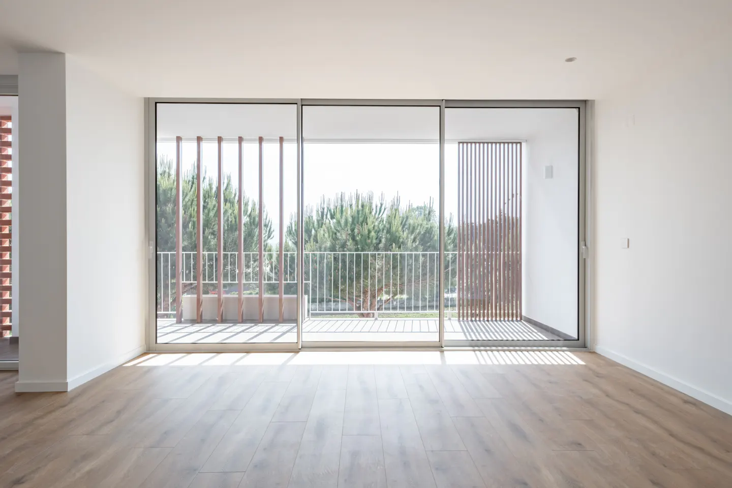 Bright, empty room with wood floors and white walls. Large sliding glass doors open to a balcony with trees in the background.