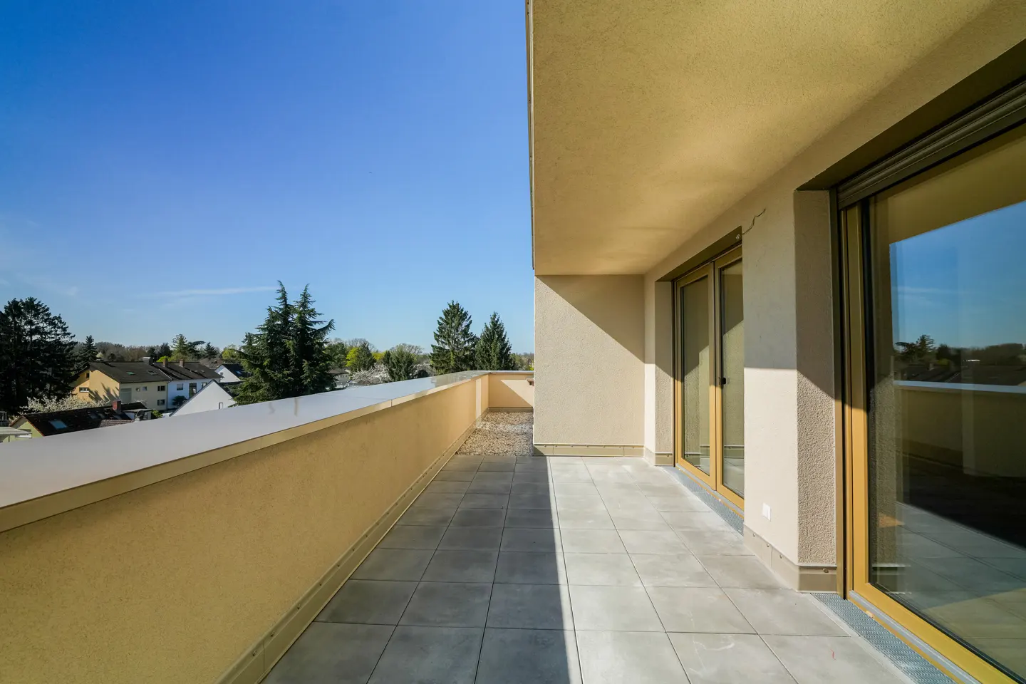 A sunny balcony with gray tiles and a beige wall overlooks trees and houses under a clear blue sky. Sliding glass doors lead inside.