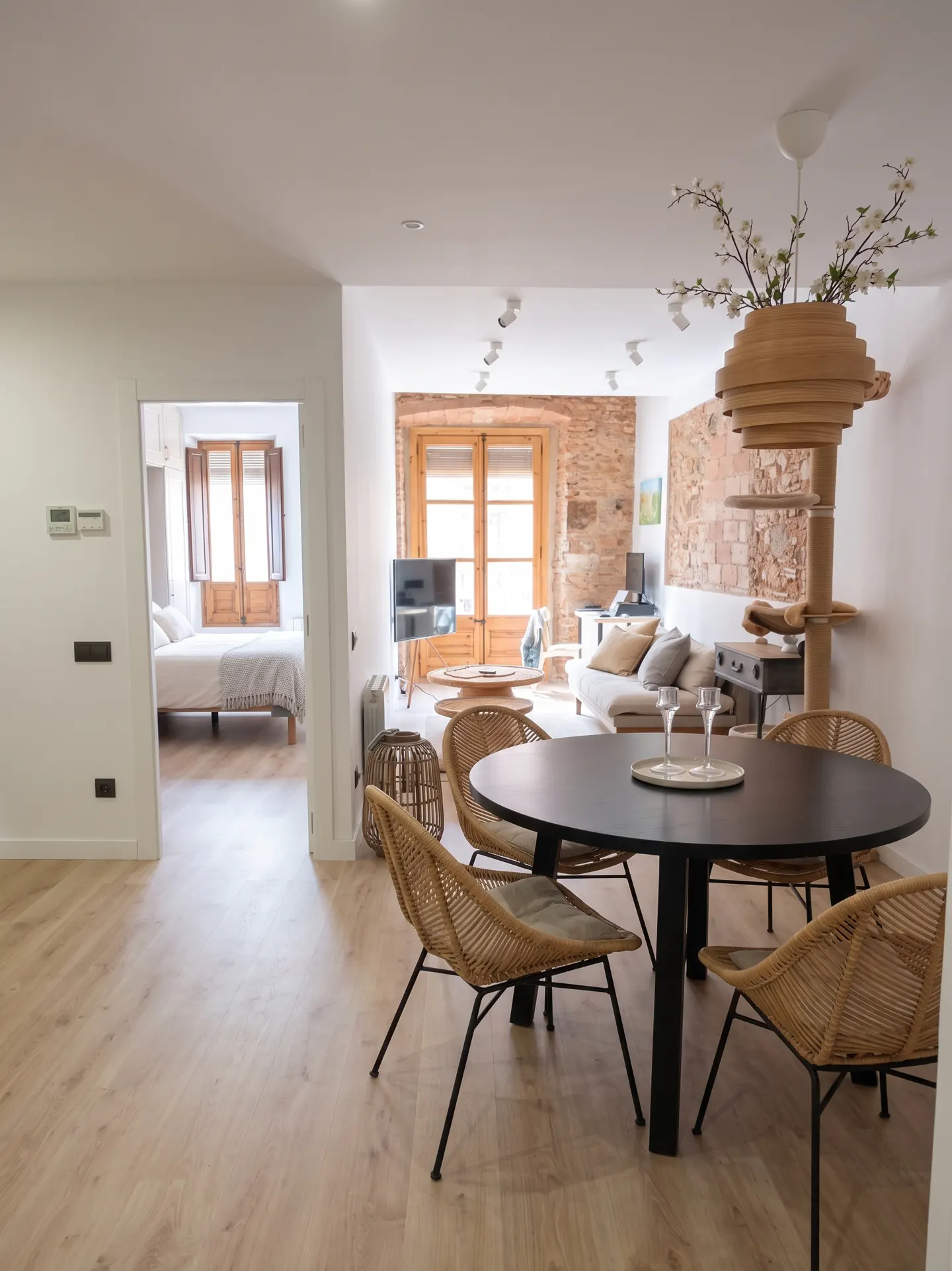 Bright apartment interior with a black round table, wicker chairs, and a view into a bedroom with a white bed.