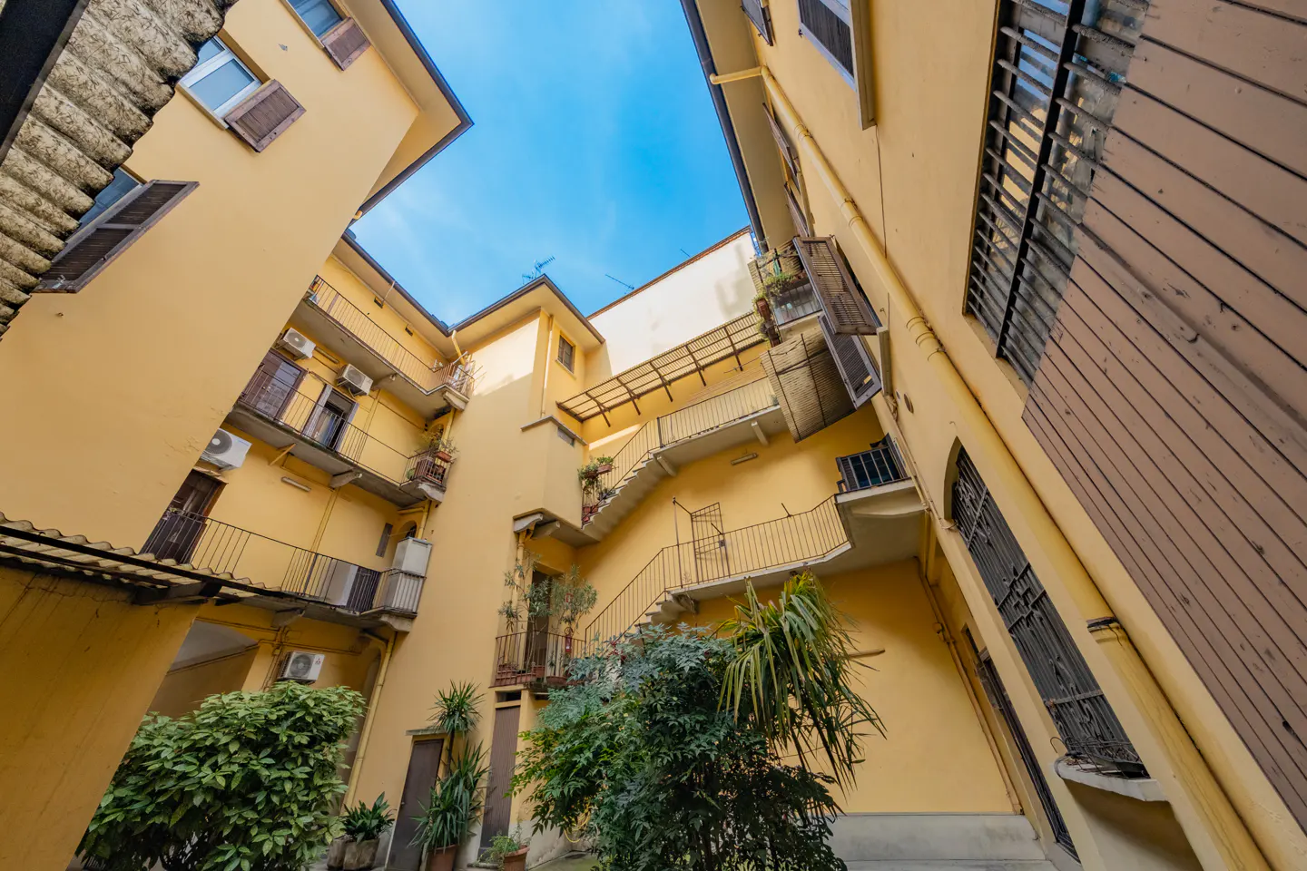 Low-angle shot of a yellow apartment building courtyard with balconies, stairs, and greenery under a blue sky.