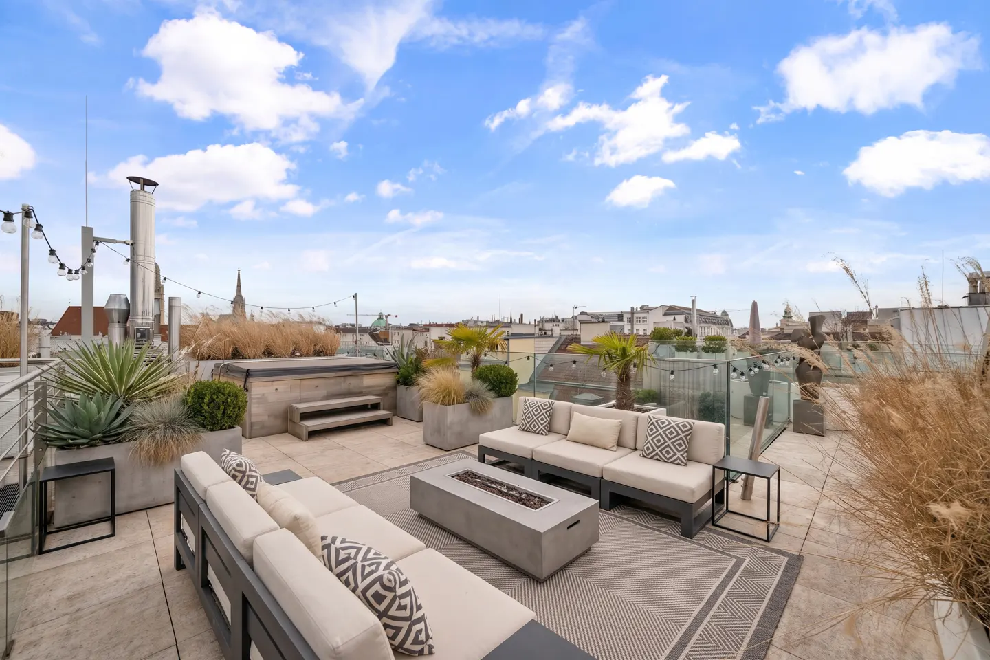 Rooftop patio with city view. Beige couches, a fire pit, and plants decorate the space under a blue sky.