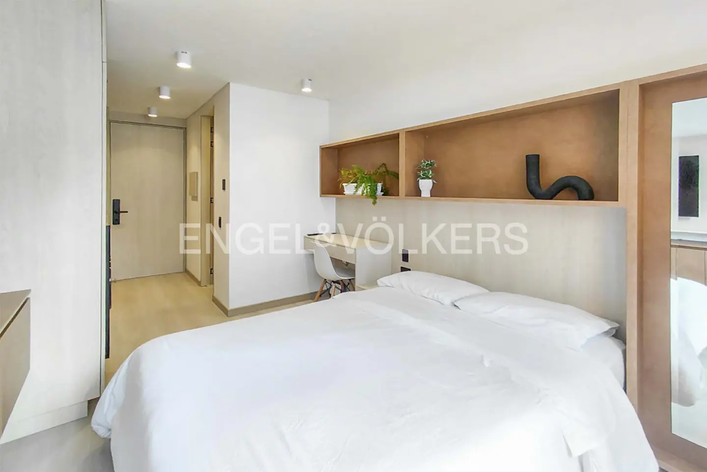 Modern bedroom with white bedding, a built-in wooden shelf, a small desk with a chair, and a hallway leading to a door.
