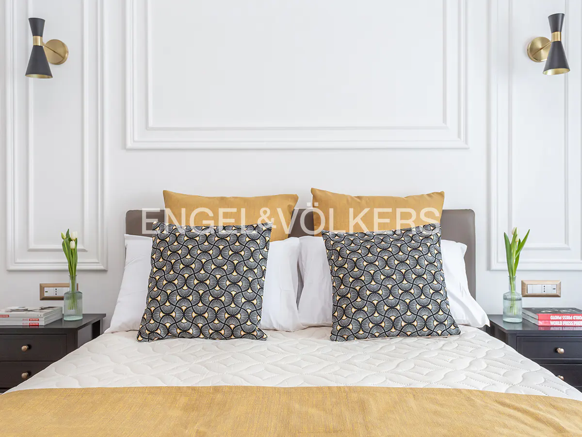 Bedroom with a bed with patterned pillows, yellow throw, and white walls with sconces.