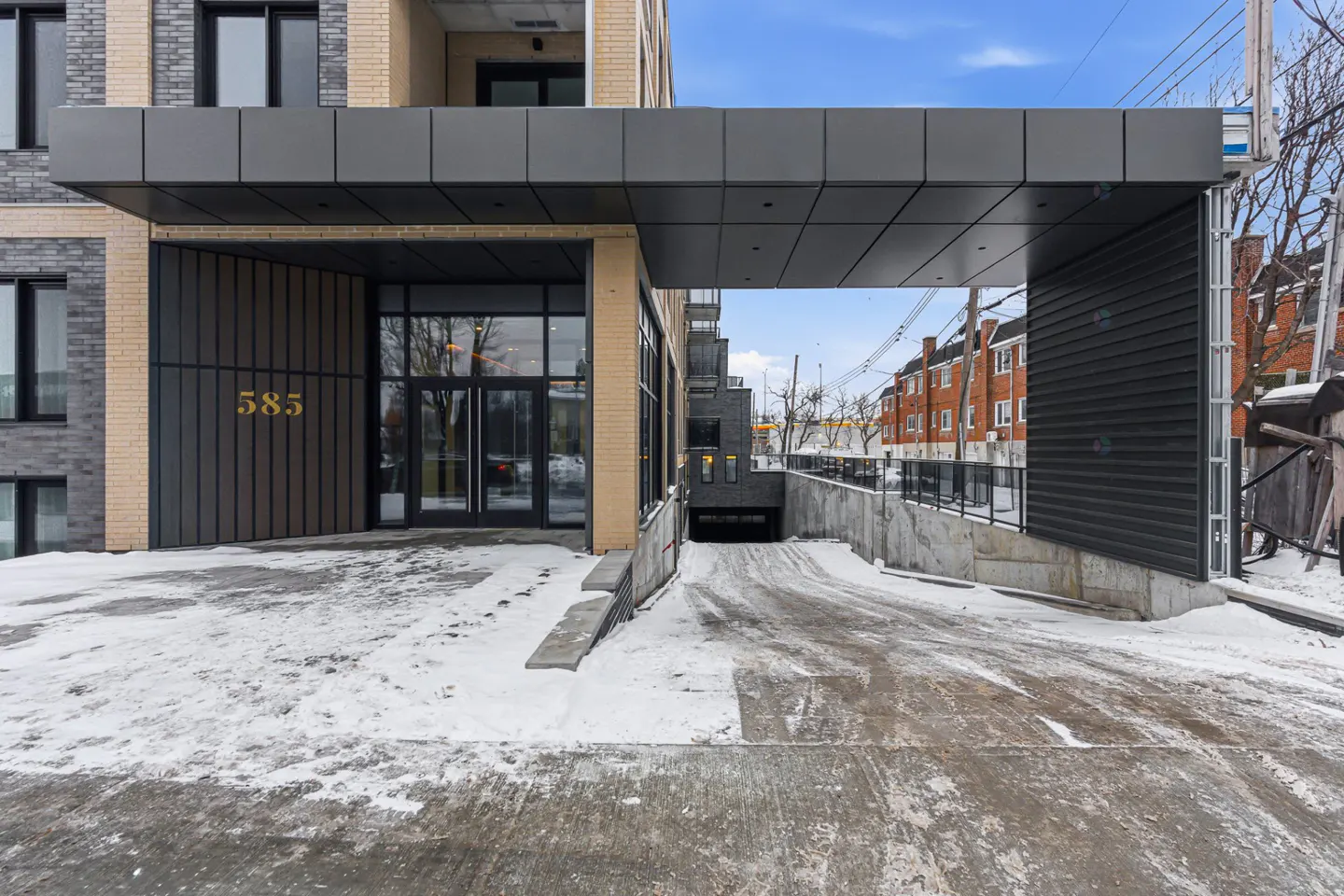 Exterior view of a modern apartment building with a covered entrance and parking garage. The building is brick and gray, with the number 585 in gold. Snow is on the ground.