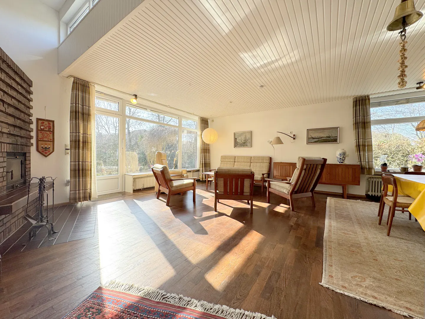 Bright living room with wood floors, a brick fireplace, and large windows. Furniture includes a sofa, chairs, and a dining table with a yellow tablecloth.