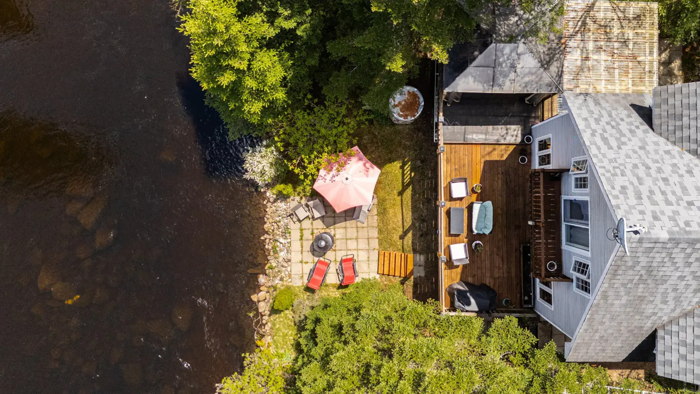 Aerial view of a gray house with a wooden deck, patio with furniture, and river access.